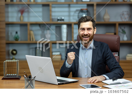Portrait of mature senior businessman in office, man looking at camera and holding hands up super power and superhero gesture, successful investor using laptop inside building. 103607502