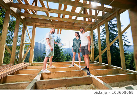 Man with plan showcases to couple the process of constructing wooden framed house. Investors inspecting their future dwelling in mountains near forest. Concept of contemporary ecological construction. 103608669