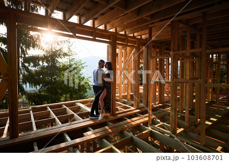 Man and woman examining their future wooden frame dwelling nestled in the mountains near forest. Youthful couple at construction site in early morning. Concept of contemporary ecological construction. Man and woman examining their future wooden frame dwelling nestled in the mountains near forest. Youthful couple at construction site in early morning. Concept of contemporary ecological construction. 103608701