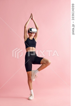 Young woman with slim body, in black sportswear and vr glasses standing in yoga pose, meditating against pink studio background 103614099