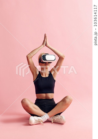 Young woman with slim body, in black sportswear and vr glasses sitting and meditating against pink studio background Young woman with slim body, in black sportswear and vr glasses sitting and meditating against pink studio background 103614117