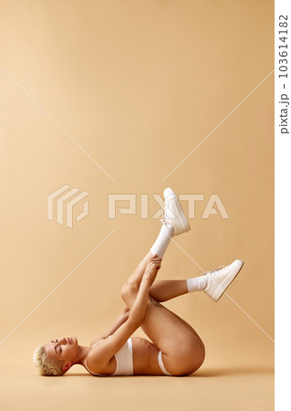 Tender, attractive young girl with blonde short hair, lying on floor in underwear and sneakers against pastel yellow studio background 103614182