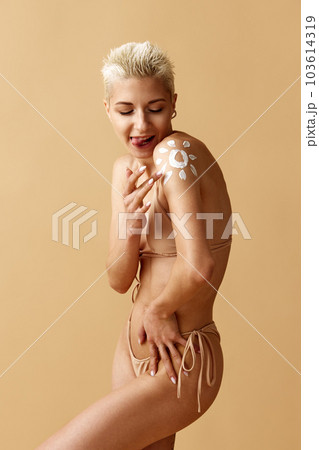 Portrait of beautiful young blonde woman applying spf cream on body, posing in swimsuit against pastel yellow studio background 103614319