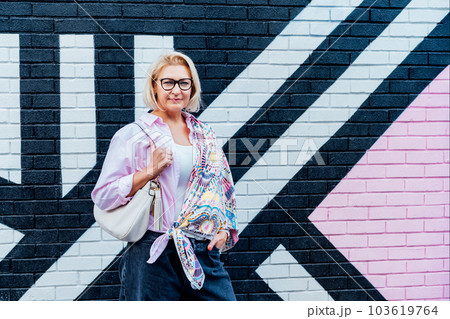 Stylish middle-aged woman wearing printed headscarf as asymmetrical cardigan posing on painted brick wall background. Creative ways to wear a headscarf in your wardrobe. Urban city street fashion. 103619764