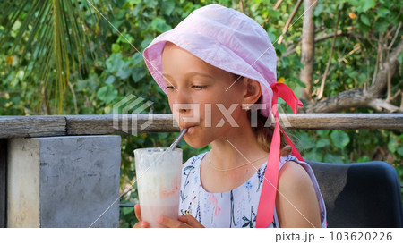 Little girl drinks strawberry milkshake outdoors, resting on a tropical terrace 103620226