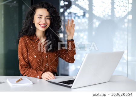 Portrait of young successful business woman in modern office using laptop, Hispanic woman with curly hair smiling and looking at camera, holding hand up in greeting gesture. 103620654