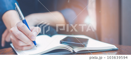 Woman hand writing on a notepad with a blue pen in the office.There is a mobile phone on the table. 103639594