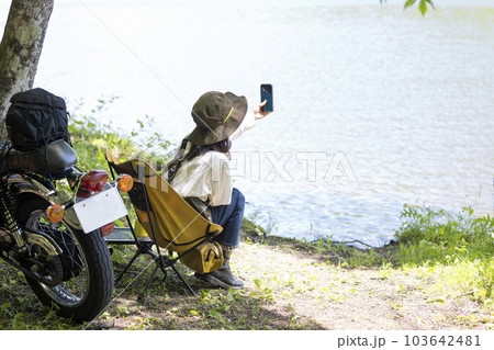 バイク女子イメージ 湖にバイクでツーリングに来てチェアリングをしながらスマホで景色を撮る女性 バイク女子イメージ 湖にバイクでツーリングに来てチェアリングをしながらスマホで景色を撮る女性 103642481