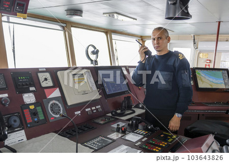 Officer on watch with radio on the navigational bridge. Caucasian man in blue uniform sweater on the bridge of cargo ship. Officer on watch with radio on the navigational bridge. Caucasian man in blue uniform sweater on the bridge of cargo ship. 103643826