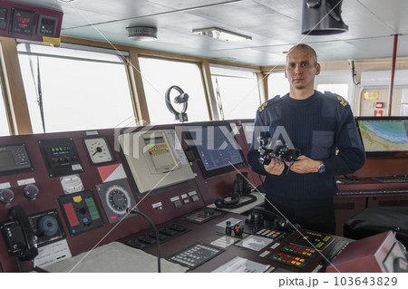 Officer on watch with binoculars on the navigational bridge. Caucasian man in blue uniform sweater on the bridge of cargo ship. Officer on watch with binoculars on the navigational bridge. Caucasian man in blue uniform sweater on the bridge of cargo ship. 103643829