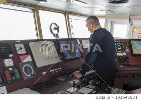 Officer on watch with radio on the navigational bridge. Caucasian man in blue uniform sweater on the bridge of cargo ship. Officer on watch with radio on the navigational bridge. Caucasian man in blue uniform sweater on the bridge of cargo ship. 103643831