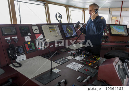 Officer on watch with radio on the navigational bridge. Caucasian man in blue uniform sweater on the bridge of cargo ship. Officer on watch with radio on the navigational bridge. Caucasian man in blue uniform sweater on the bridge of cargo ship. 103643834