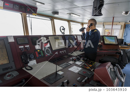 Officer on watch with radio and binoculars on the navigational bridge. Caucasian man in blue uniform sweater on the bridge of cargo ship. Officer on watch with radio and binoculars on the navigational bridge. Caucasian man in blue uniform sweater on the bridge of cargo ship. 103643910