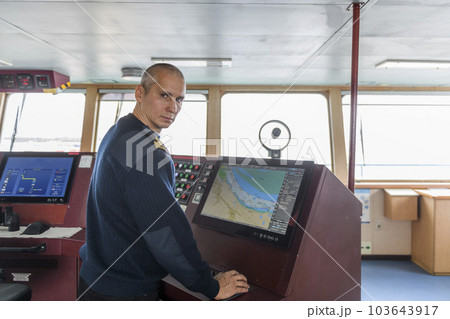 Officer on watch with electronic chart on the navigational bridge. Caucasian man in blue uniform sweater using ECDIS on the bridge of cargo ship. Officer on watch with electronic chart on the navigational bridge. Caucasian man in blue uniform sweater using ECDIS on the bridge of cargo ship. 103643917