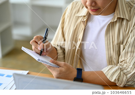 Young businessman looking at laptop screen and making important notes, planning daily appointment in diary Young businessman looking at laptop screen and making important notes, planning daily appointment in diary 103646655