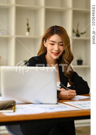 Portrait of beautiful female manager in trendy suit using laptop and making notes on notebook 103646681
