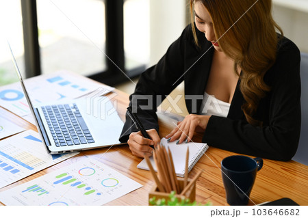Businesswoman writing notes in personal daily planner at her office desk with laptop and financial document 103646682