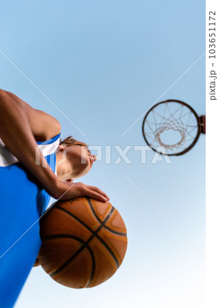 Basketball. A teenager in blue sportswear holds a ball in his hands and looks at a basketball Hoop. Bottom view. Sky in the background. Concept of sports games Basketball. A teenager in blue sportswear holds a ball in his hands and looks at a basketball Hoop. Bottom view. Sky in the background. Concept of sports games 103651172