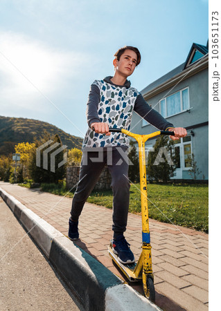 A teenage boy with a serious face rides a scooter on the sidewalk. In the background, an empty street. Bottom view. Copy space. Concept of sports recreation and youth activity 103651173