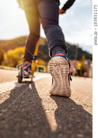 A teenage boy rides a scooter. Right foot in a sneaker close-up. Bottom and rear view. Empty street in Sunny weather in the background. Concept of youth activity, sports and recreation 103651175