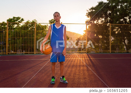 Basketball. A teenage boy in blue sportswear stand and poses holding a basketball in his hand. In the background there is a sports field and a sunset. Concept of sports games 103651205