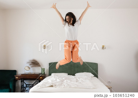Happy young Caucasian woman dances and jumps on bed in bedroom. Freedom and relax at home Happy young Caucasian woman dances and jumps on bed in bedroom. Freedom and relax at home 103651240