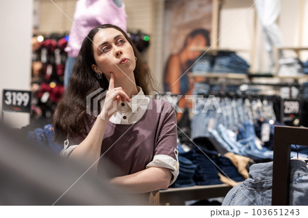 Portrait of thoughtful Caucasian woman looking at hangers with clothes in store. Shopping in mall. Sale and purchase Portrait of thoughtful Caucasian woman looking at hangers with clothes in store. Shopping in mall. Sale and purchase 103651243