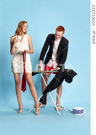 Portrait of happy young couple getting ready to go out against blue studio background. Woman drinking coffee, man ironing his pants 103651325