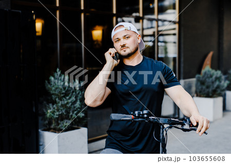 Caucasian young handsome man standing on street at electric scooter and talking on mobile phone. Stylish guy speaking on cellphone outdoors 103655608