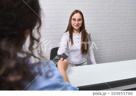 Smiling pleasant receptionist accepting cashless payment, holding out pos terminal to a client customer for payment using credit card and NFC technology. People. Customer service. Technology. Finance 103655780