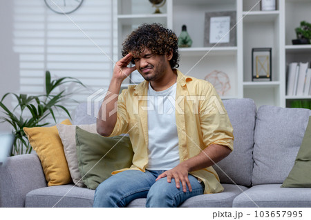 Young man sitting sick alone at home in living room on sofa, Hispanic man has severe headache and migraine. Young man sitting sick alone at home in living room on sofa, Hispanic man has severe headache and migraine. 103657995