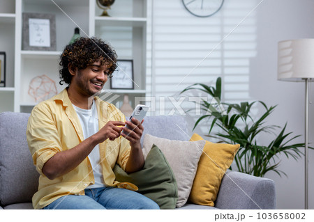 A young Hispanic man is sitting at home on the sofa in the living room, the man is holding a phone in his hands, using an online application on a smartphone, browsing the Internet and typing messages. A young Hispanic man is sitting at home on the sofa in the living room, the man is holding a phone in his hands, using an online application on a smartphone, browsing the Internet and typing messages. 103658002