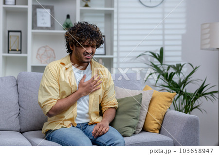 Sick man having severe chest pain and heart attack, hispanic young man sitting alone at home in living room, holding hands to chest. 103658947