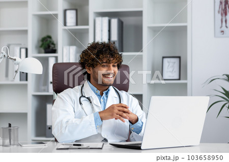 Young successful doctor working inside hospital office, man smiling and looking at laptop screen, hispanic man in medical gown is satisfied with patient's treatment achievement results. Young successful doctor working inside hospital office, man smiling and looking at laptop screen, hispanic man in medical gown is satisfied with patient's treatment achievement results. 103658950