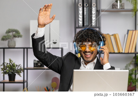 Indian businessman wearing headphones listening music dancing after working on laptop at home office 103660367