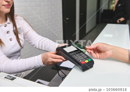 Close-up view of customer's hand paying by credit card and entering pin code on reader holded by a pleasant female manager administrator in the reception desk of modern medical clinic. NFC technology. Close-up view of customer's hand paying by credit card and entering pin code on reader holded by a pleasant female manager administrator in the reception desk of modern medical clinic. NFC technology. 103660918