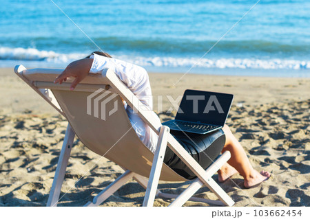 Back view of alone young brunette woman freelancer in office clothes with a laptop looking on the sea sitting on the deck chair on beach. Summer concept Back view of alone young brunette woman freelancer in office clothes with a laptop looking on the sea sitting on the deck chair on beach. Summer concept 103662454
