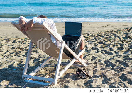 Back view of alone young brunette woman freelancer in office clothes with a laptop looking on the sea sitting on the deck chair on beach. Summer concept 103662456
