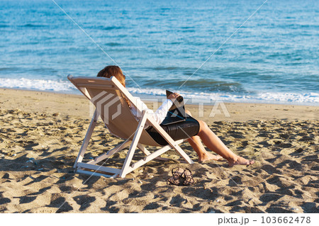 Back view of alone young brunette woman freelancer in office clothes with a laptop looking on the sea sitting on the deck chair on beach. Summer concept 103662478
