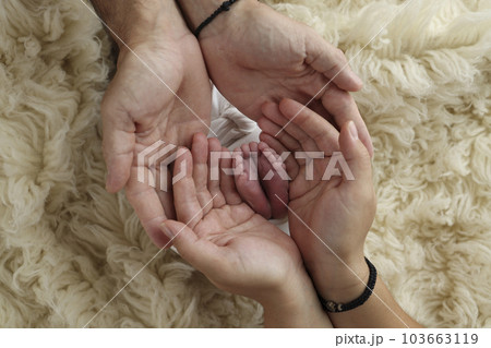 Feet of the newborn on the palms of the parents. The palms of the father and mother are holding the foot of the newborn baby in a white flokati blanket. Photography of a child's toes, heels and feet. 103663119