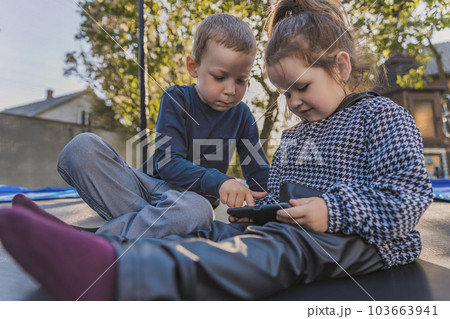 children sit on a trampoline and look at the phone children sit on a trampoline and look at the phone 103663941