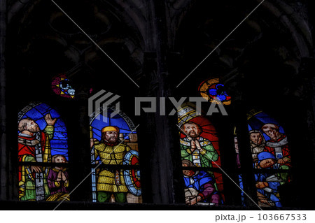 Multi colored medieval era stained glass panels depicting clergy, soldiers, saints set in stone gothic tracery in a cathedral. Illuminated windows against a dark background. Vivid colors 103667533