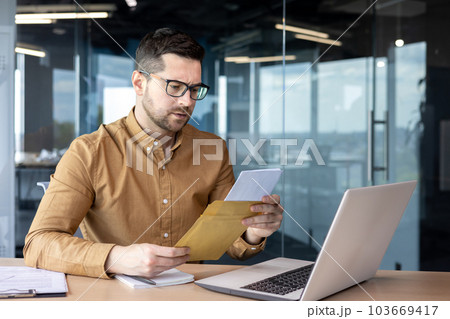 A serious and worried young businessman working in the office holds a letter, documents. got bad news. Saddenedly reads a message, a contract, an agreement. 103669417