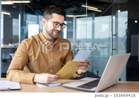 A young man businessman sits smiling in the office at the table and holds in his hands and reads documents, a contract, a successful deal that he received by mail. 103669418