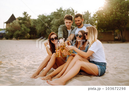 Group of friends cheers and drink beers on the beach. Young friends relaxing and having picnic. Suummertime. 103672298