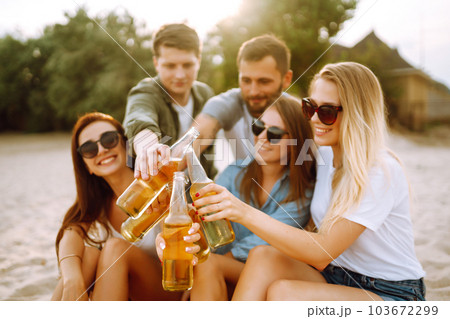 Group of friends cheers and drink beers on the beach. Young friends relaxing and having picnic. Suummertime. 103672299