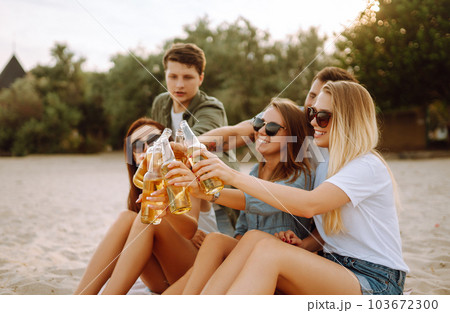 Group of friends cheers and drink beers on the beach. Young friends relaxing and having picnic. Suummertime. 103672300