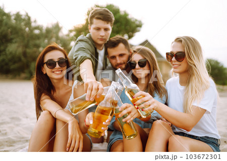 Group of friends cheers and drink beers on the beach. Young friends relaxing and having picnic. Suummertime. 103672301