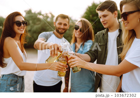 Group of friends cheers and drink beers on the beach. Young friends relaxing and having picnic. Suummertime. 103672302