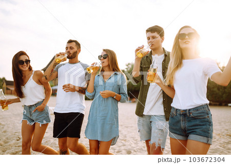 Group of friends cheers and drink beers on the beach. Young friends relaxing and having picnic. Suummertime. 103672304